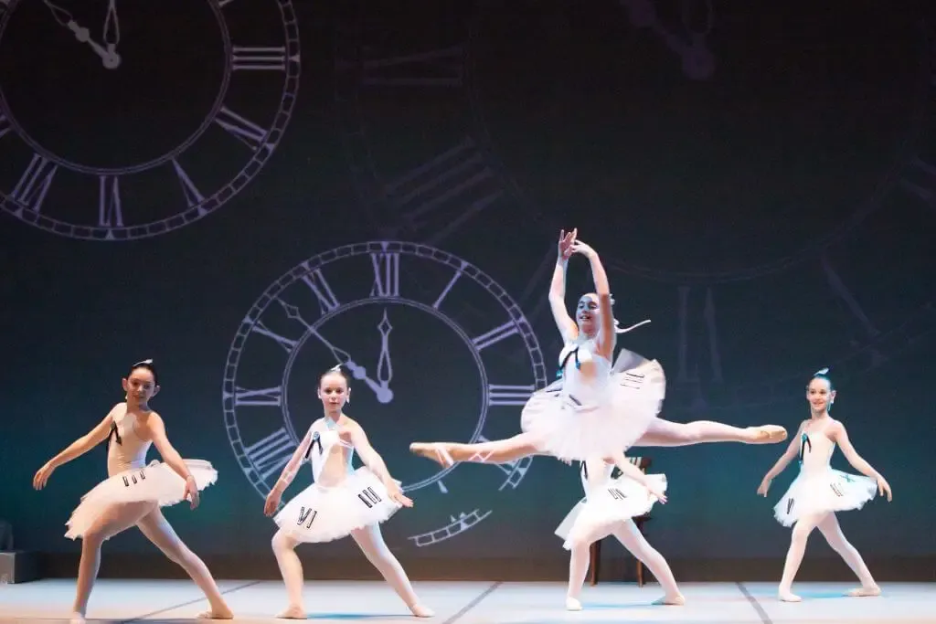 Allieve di danza classica in tutù sul palco, con un grande orologio sullo sfondo